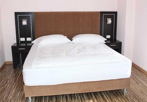 A neatly made bed with a brown headboard, two bedside tables with lamps, and a striped carpet. - Nais Beach Durres (Durrës)