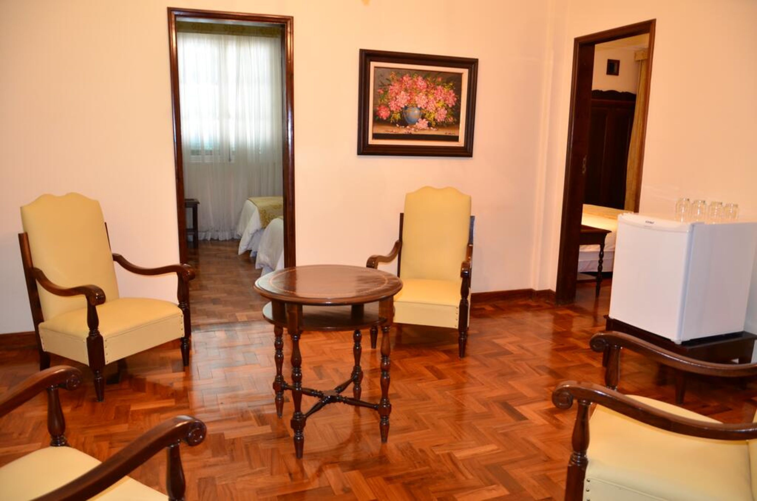 A room with wooden flooring, two armchairs, a round table, and a refrigerator.