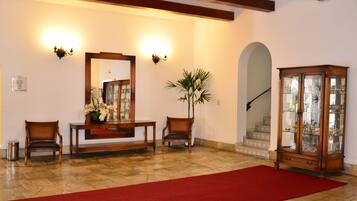 Un área de recepción con alfombra roja, sillas de madera, una consola con flores, apliques, un espejo grande y un vitrina de vidrio.