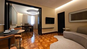 A hotel room with a wooden parquet floor, a dining area with a table and chairs, a sofa, a TV mounted on the wall, and a door leading to another room. - Glória Caxambu Resort & Convention (Caxambu)