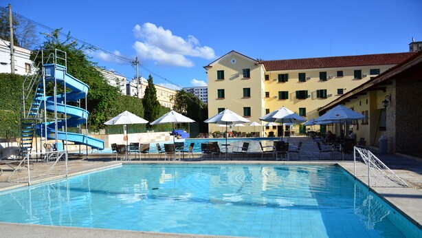 A swimming pool with a blue slide, lounge chairs, umbrellas, and a building with a red roof in the background. - Glória Caxambu Resort & Convention (Caxambu)