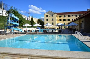 A swimming pool with a blue slide, lounge chairs, umbrellas, and a building with a red roof in the background. - Glória Caxambu Resort & Convention (Caxambu)