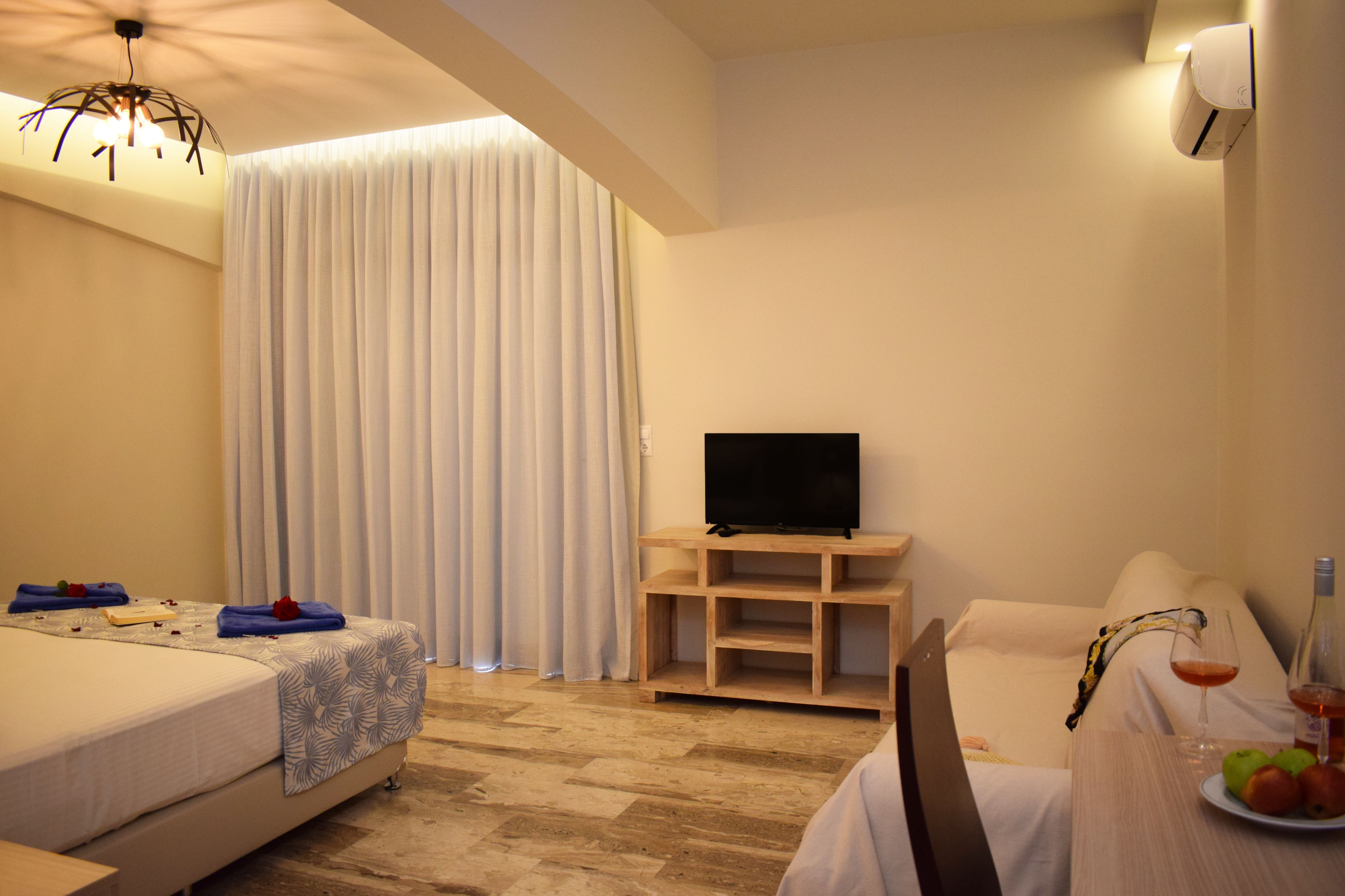 A hotel room with a bed, a TV on a wooden stand, a dining table with a plate of fruit and a glass of wine, and a large window with curtains.