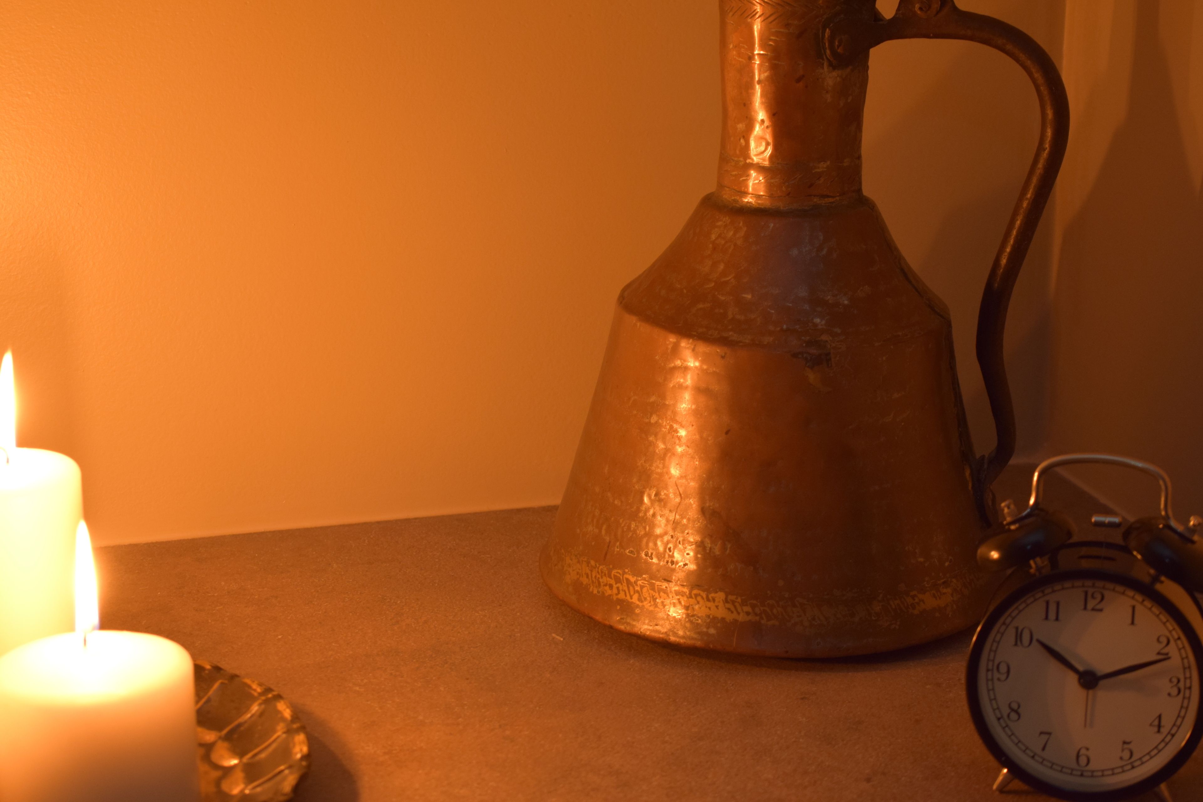 A metal pitcher, a lit candle, and an alarm clock on a table.