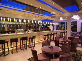 Un área de bar con una barra alargada, varios taburetes y mesas redondas con sillas.