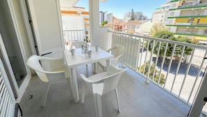 Un balcón con mesa y sillas, una puerta blanca y vistas a edificios al fondo.