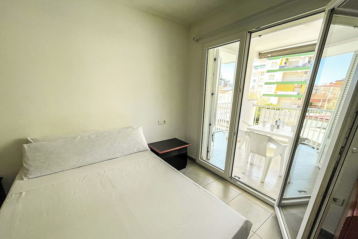 A bedroom with a bed, a small table, and a sliding glass door leading to a balcony with a view of buildings.