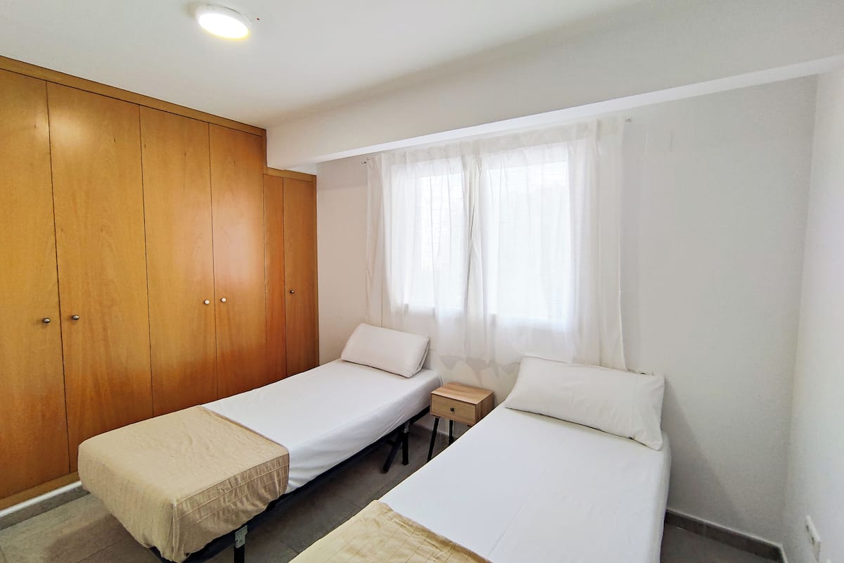 A small, clean room with two single beds, a wooden wardrobe, a small bedside table, and a window with sheer curtains.
