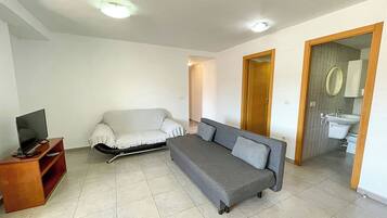 Un salon avec un canapé, un téléviseur posé sur un meuble en bois et une porte donnant sur une salle de bain.
