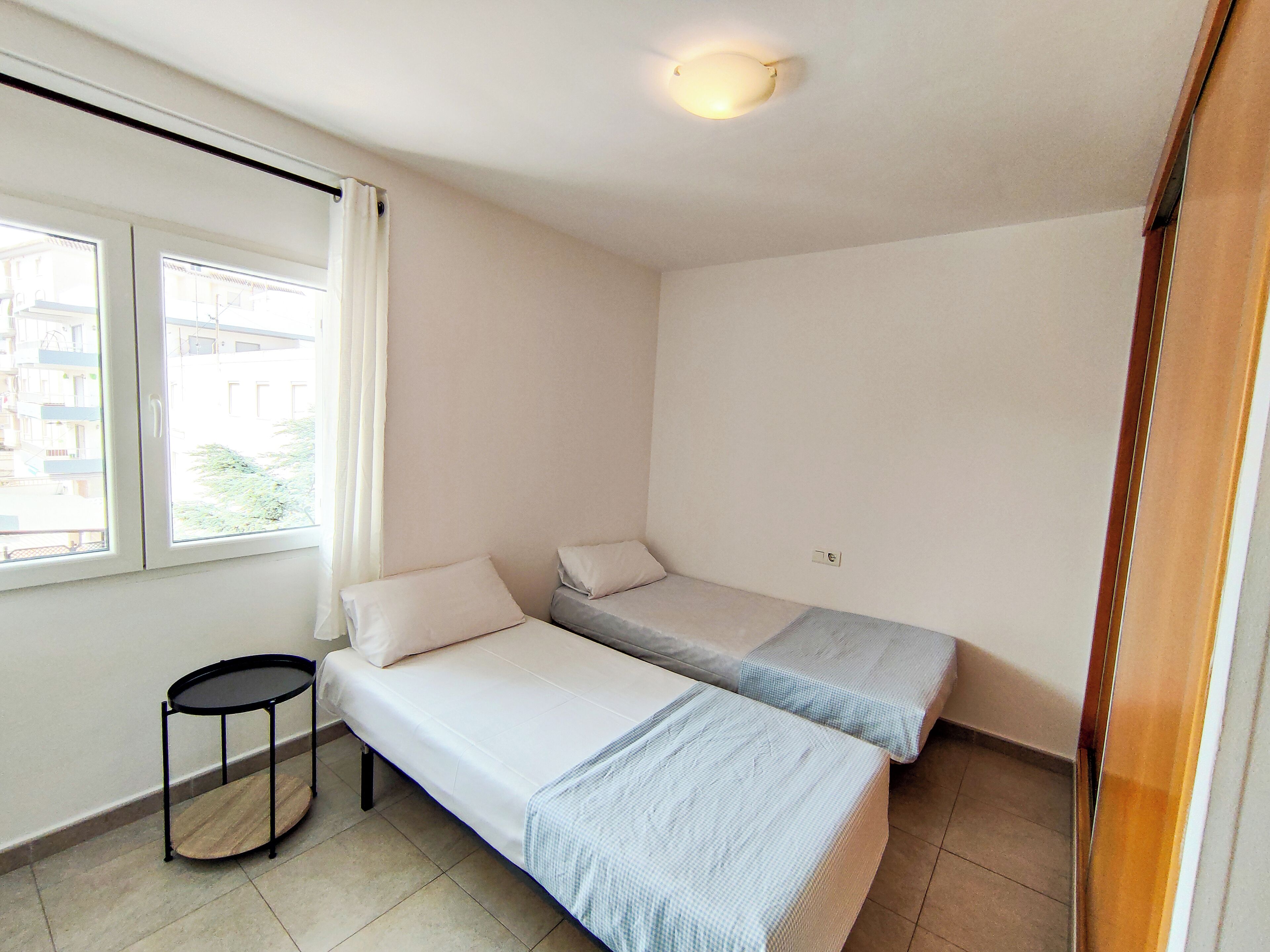 Una habitación pequeña y luminosa con dos camas individuales, una mesita y una ventana con cortinas.