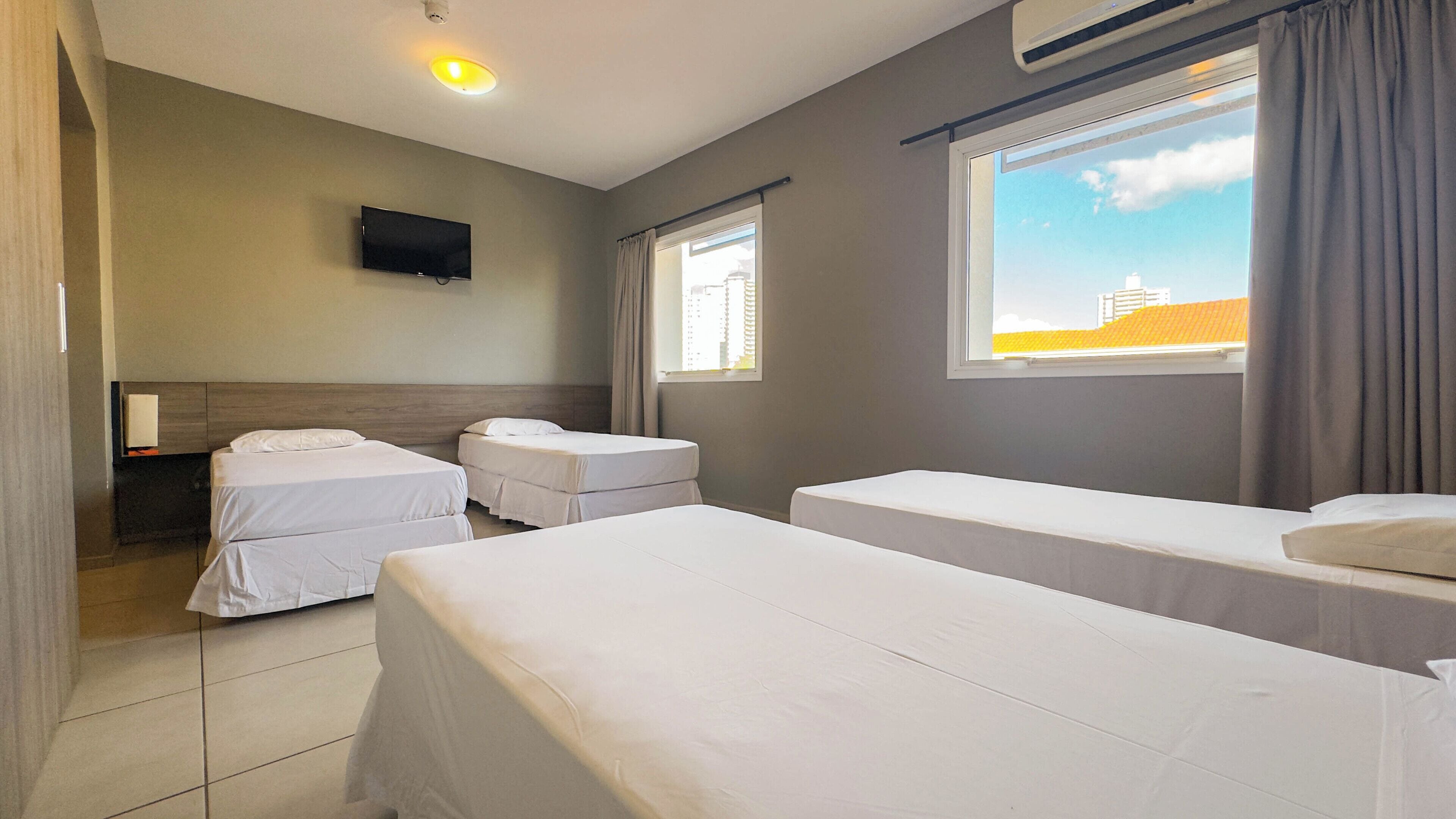 Quarto de hotel com três camas de solteiro, uma TV de tela plana na parede e uma janela com cortinas.
