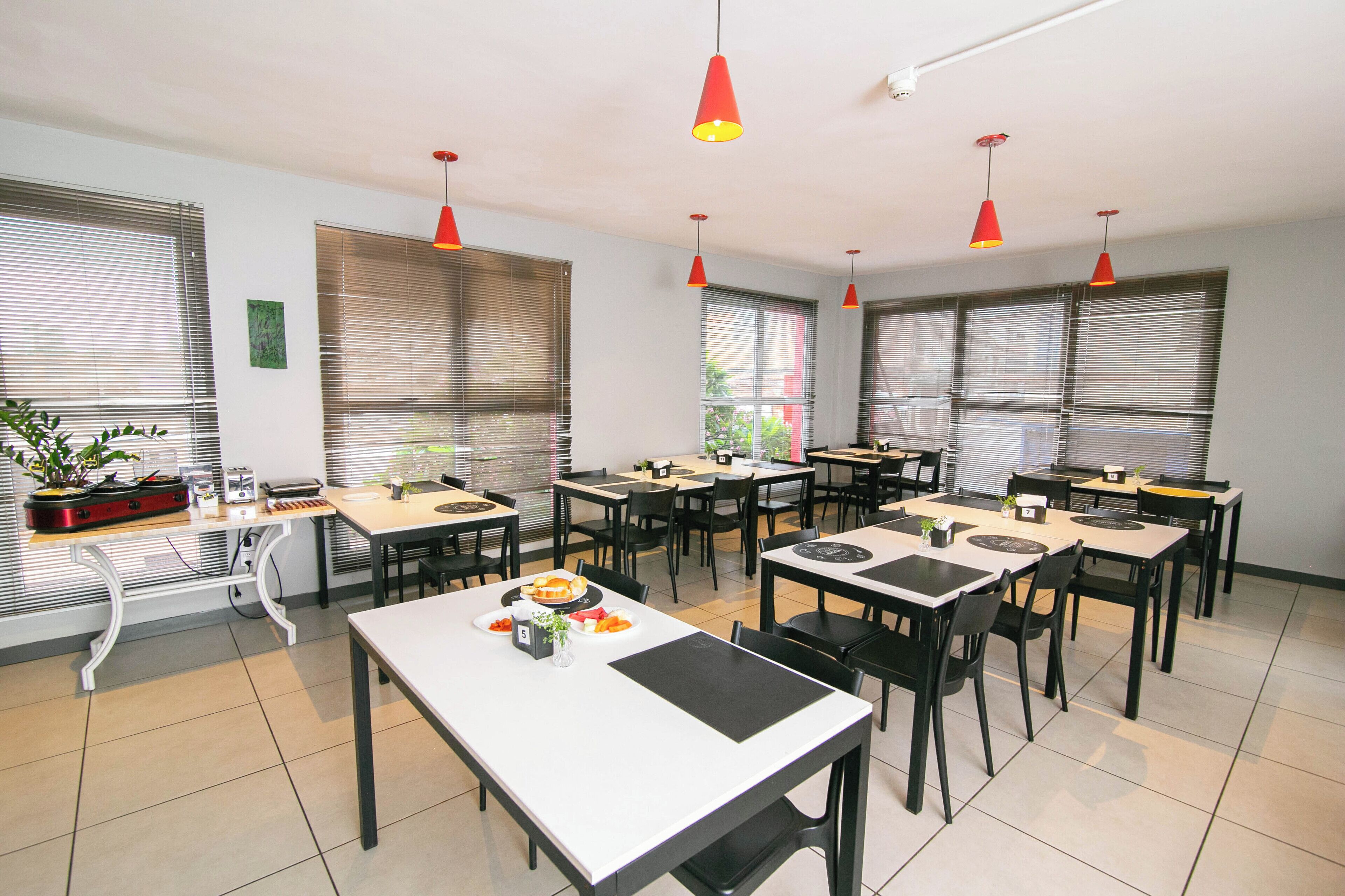 Un espace repas comprenant plusieurs tables et chaises, des suspensions rouges et de grandes fenêtres équipées de stores.