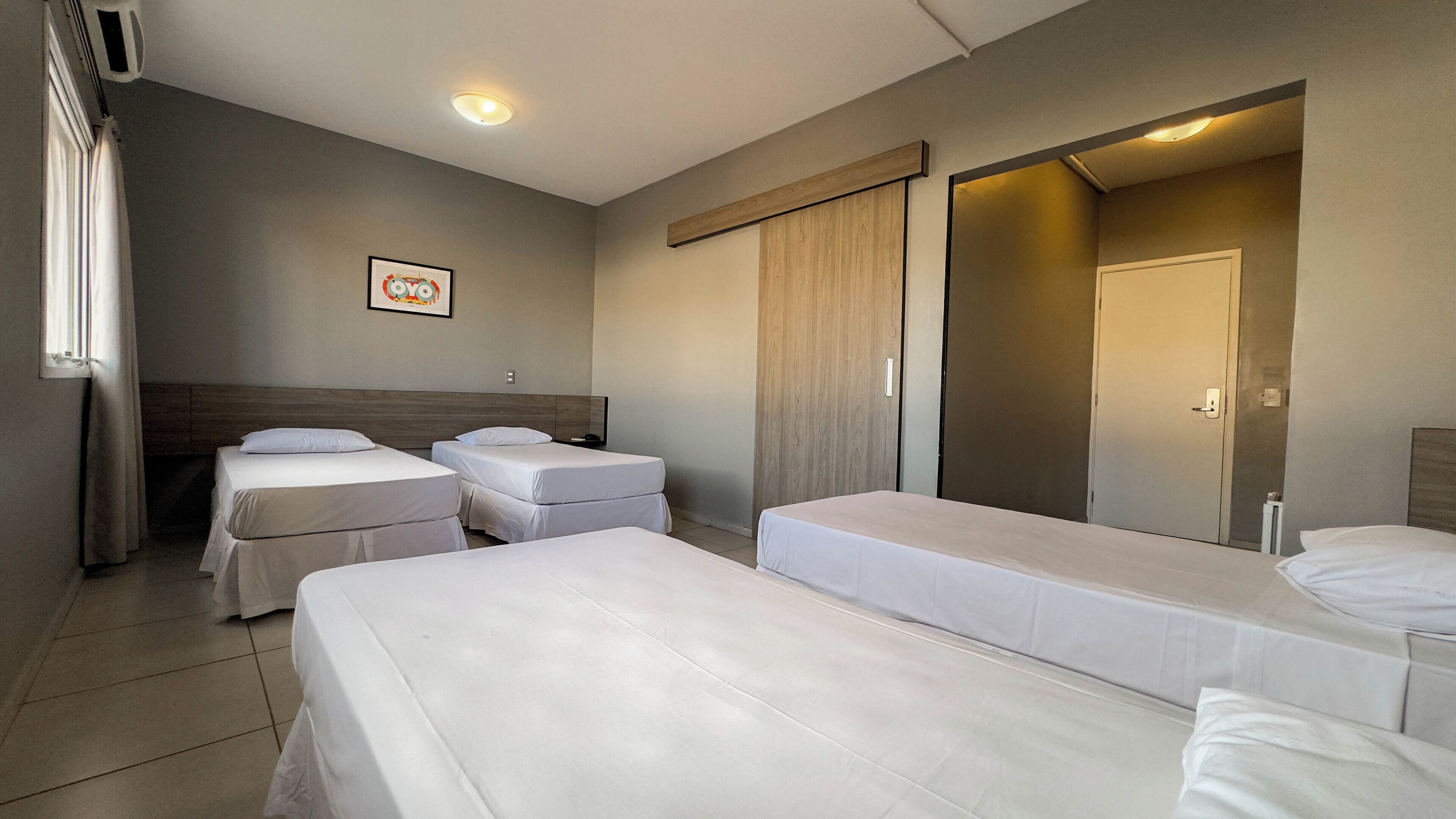 Habitación de hotel con cuatro camas individuales, un espejo grande, cabecera de madera, ventana con cortinas e imagen colgada en la pared.