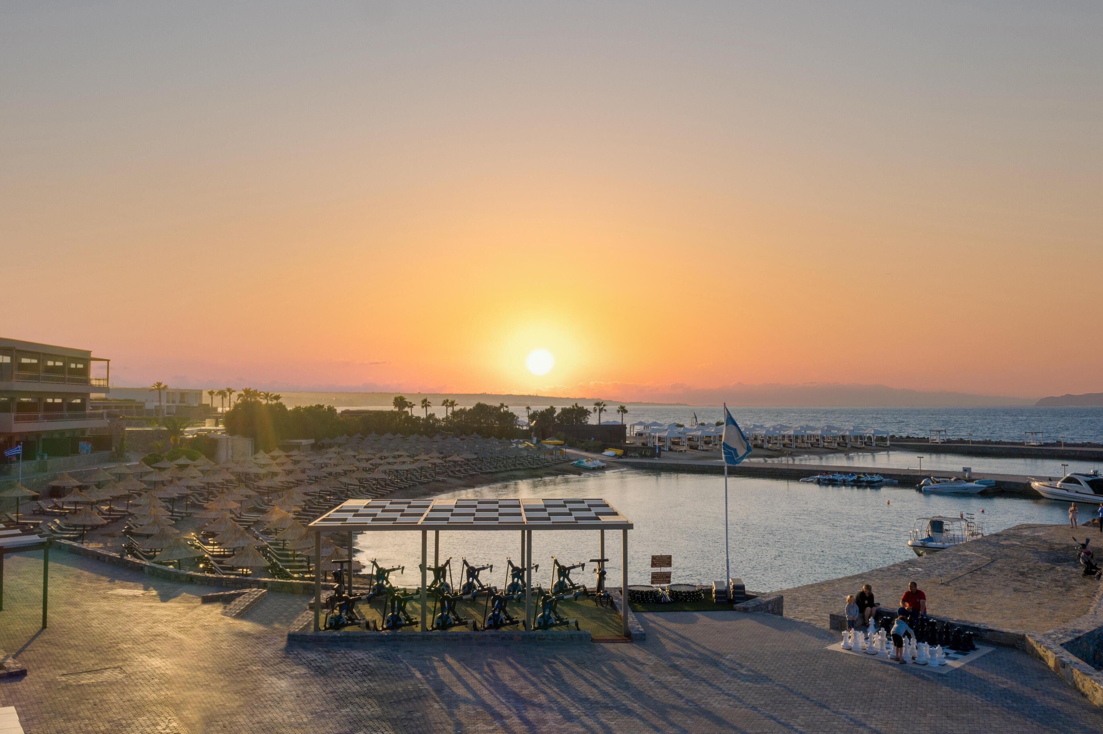 Vista à beira-mar com pôr do sol, espaço coberto para bicicletas e barcos na água.
