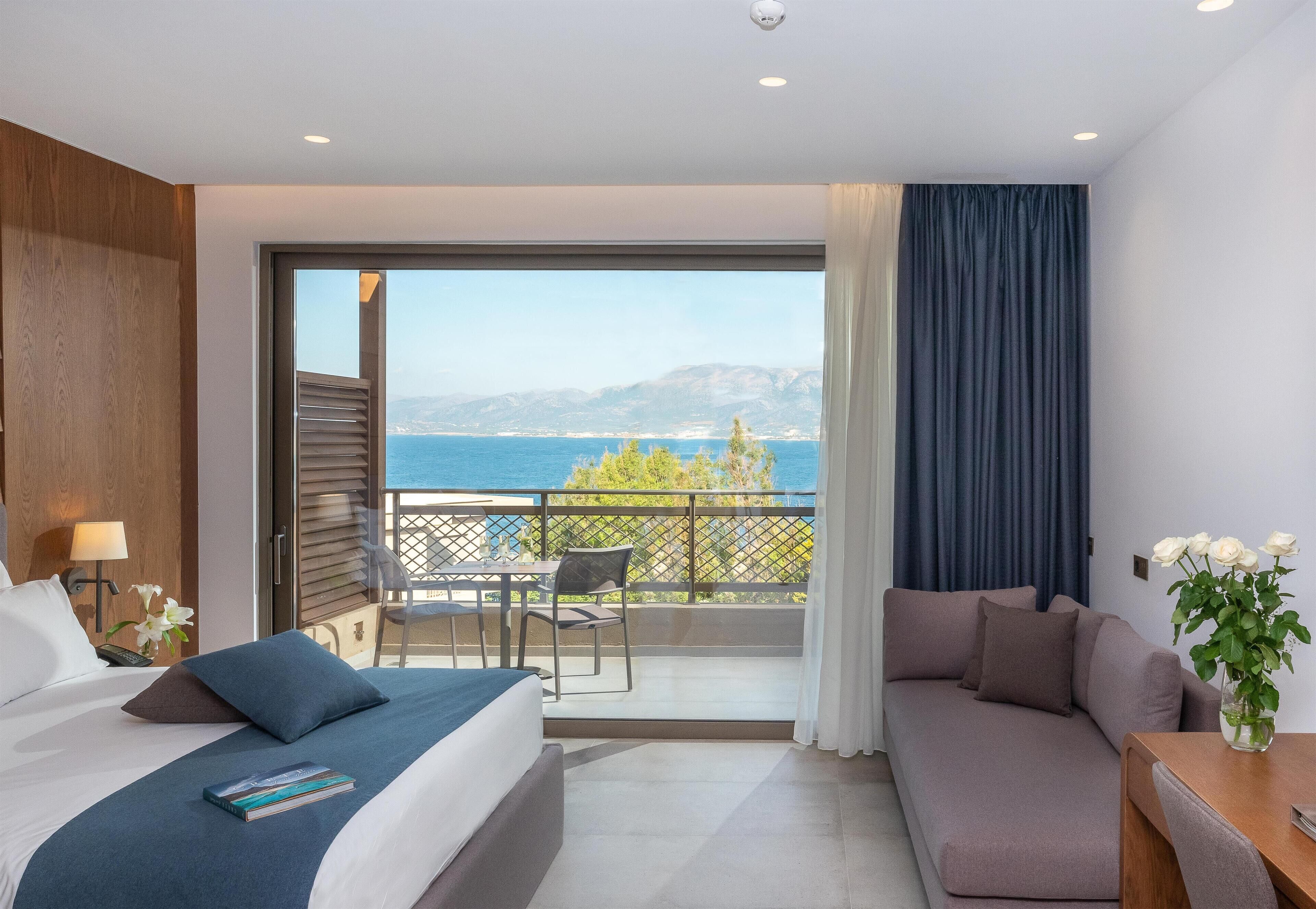 A hotel room with a bed, a sofa, a balcony with a view of the sea, and a dining area.