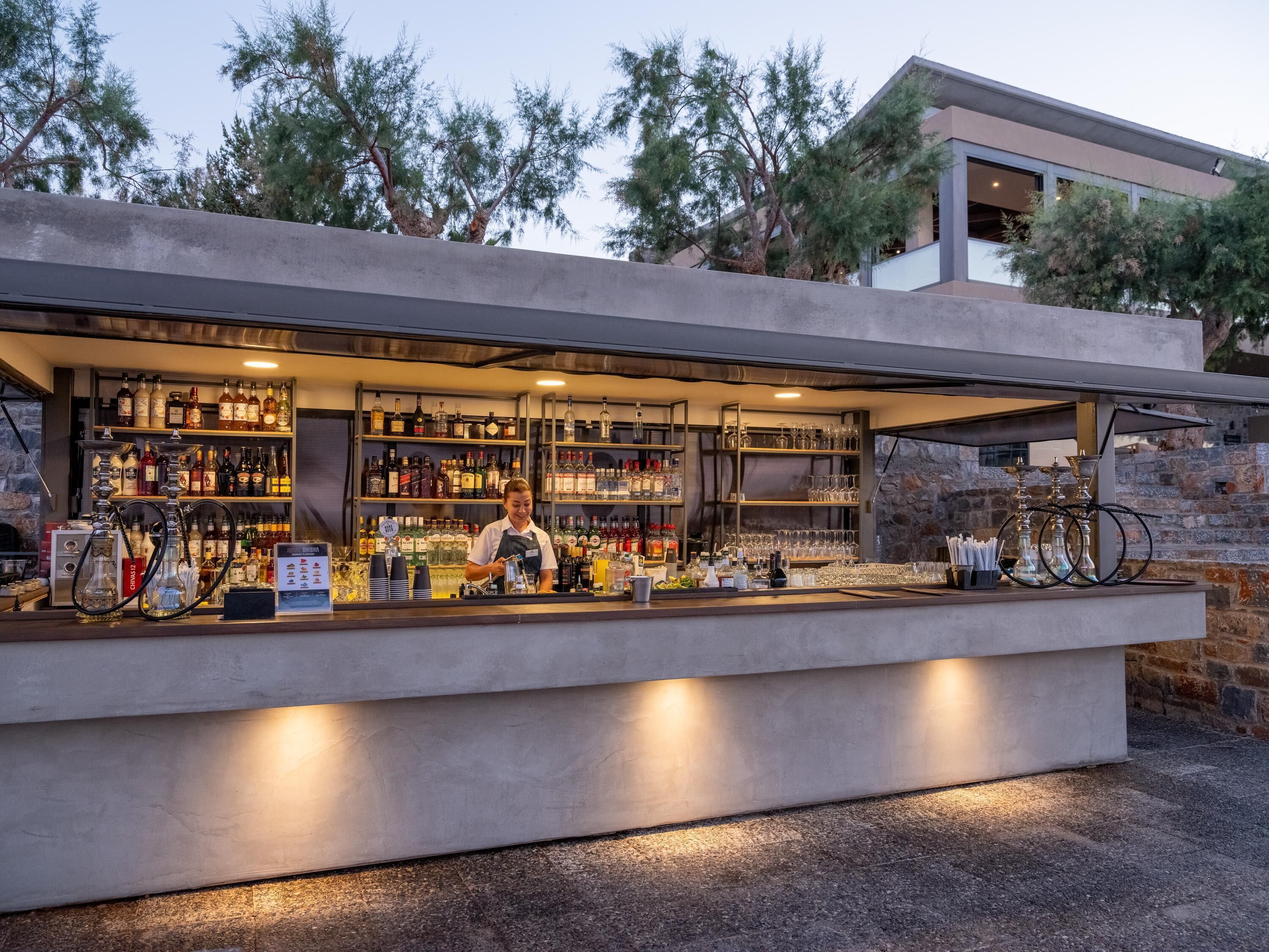 A modern bar with a bartender, shelves of liquor, and a stone wall backdrop.