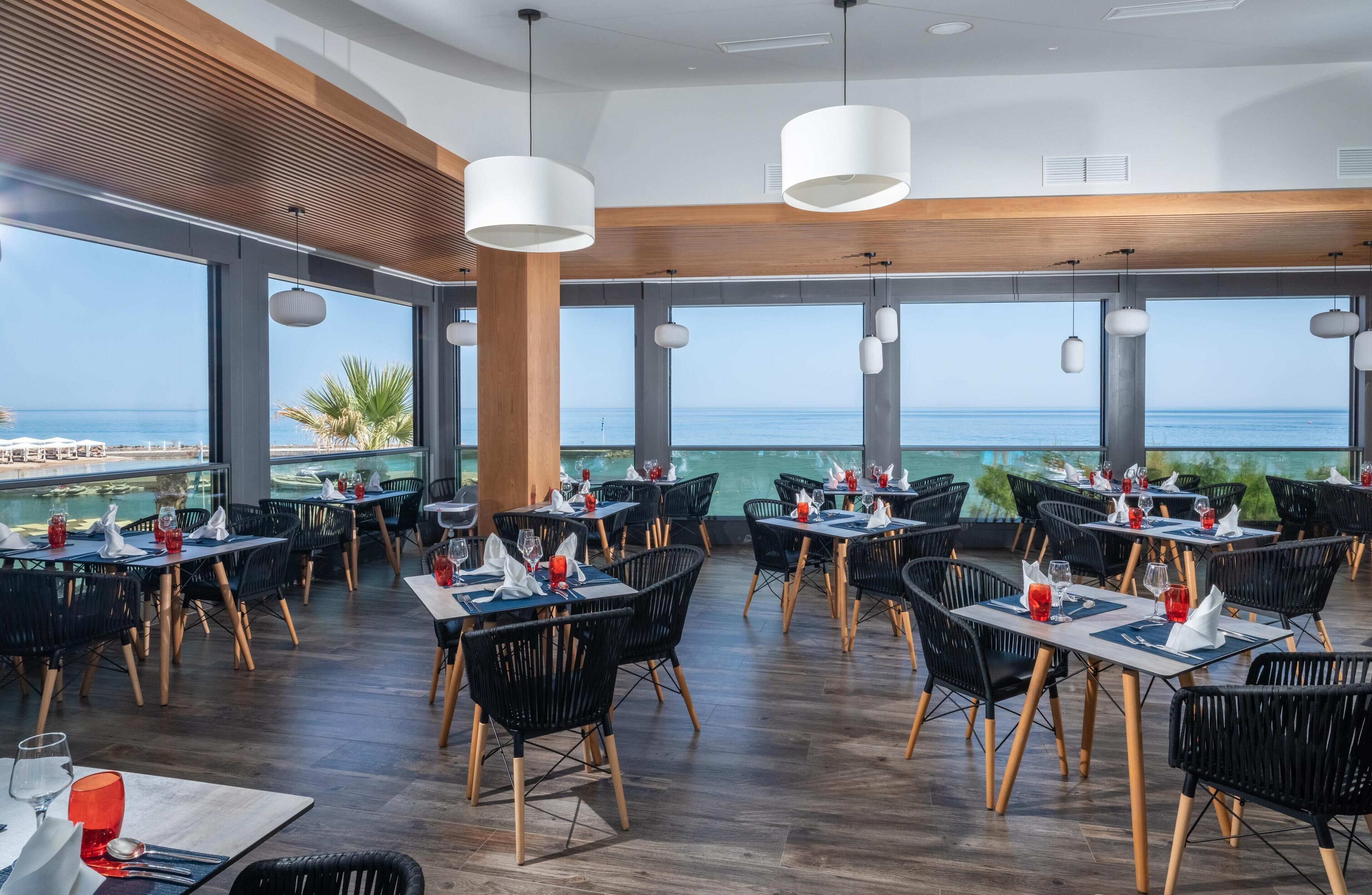 Un restaurant avec des tables et des chaises disposées pour accueillir les clients, de grandes fenêtres offrant une vue sur la mer et des lampes suspendues décoratives.