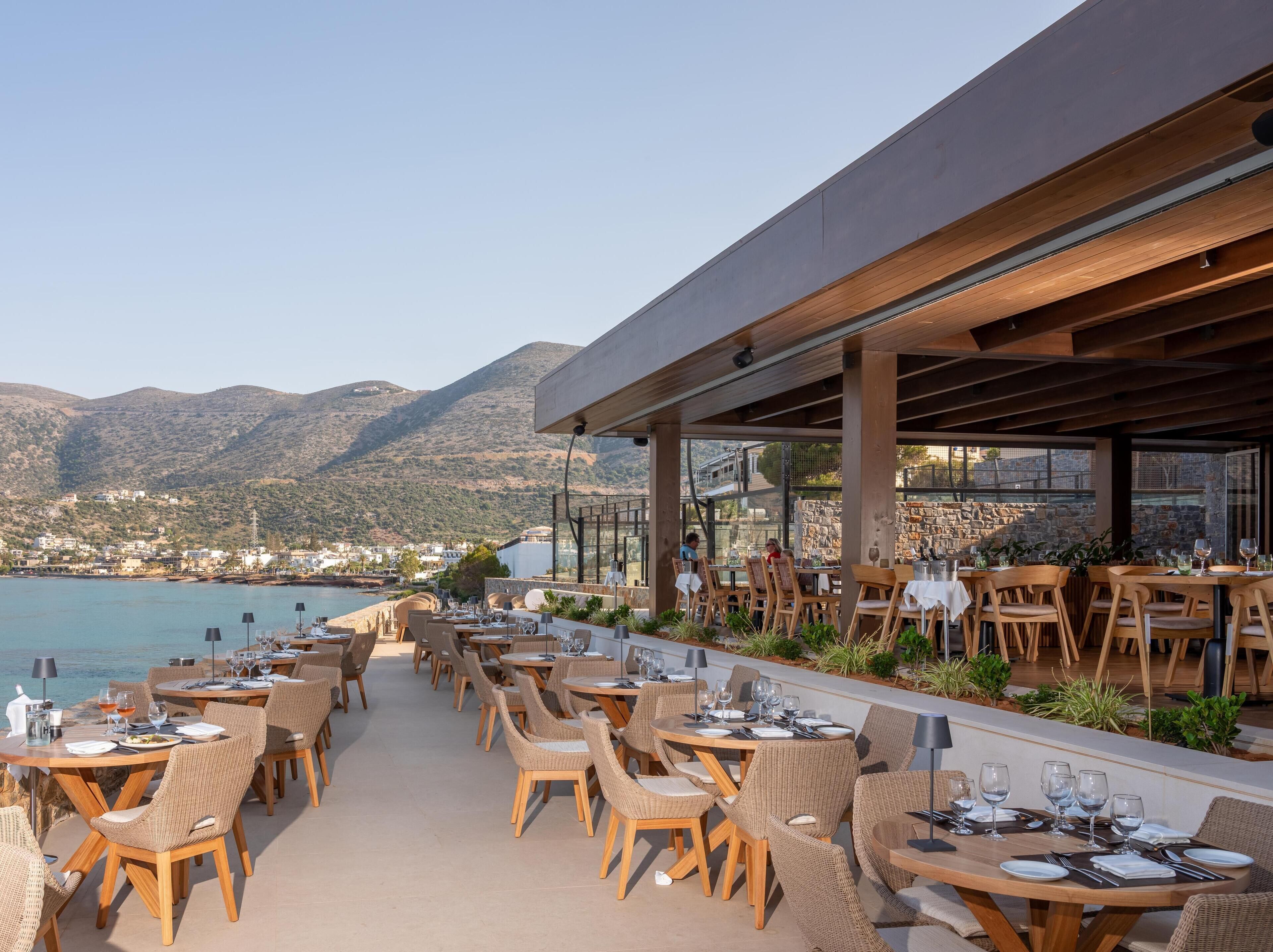 An outdoor dining area with tables and chairs, overlooking a body of water and hills.