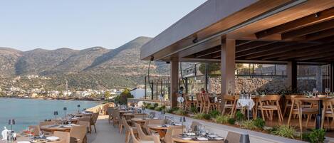 An outdoor dining area with tables and chairs, overlooking a body of water and hills.