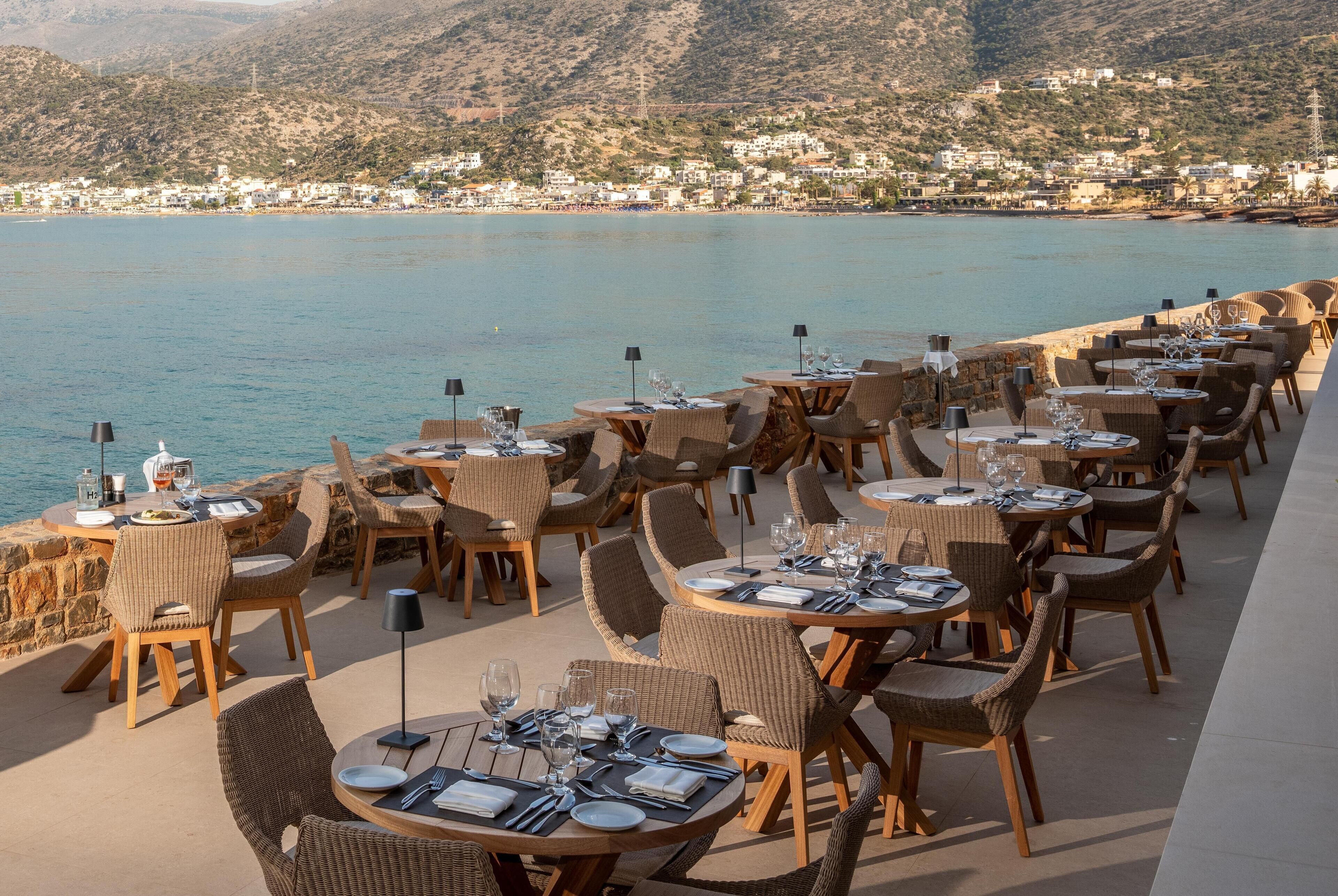 A seaside restaurant with round tables and wicker chairs, overlooking a calm sea and a hillside town.