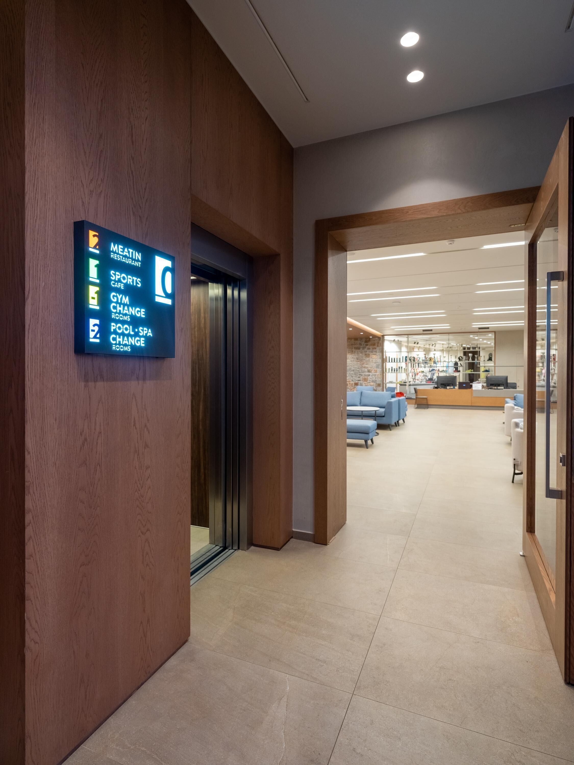 Un hall d'hôtel comprenant un comptoir d'accueil, un espace salon et un panneau indiquant les différents services proposés, tels qu'un restaurant, un café sportif, une salle de sport et un spa.