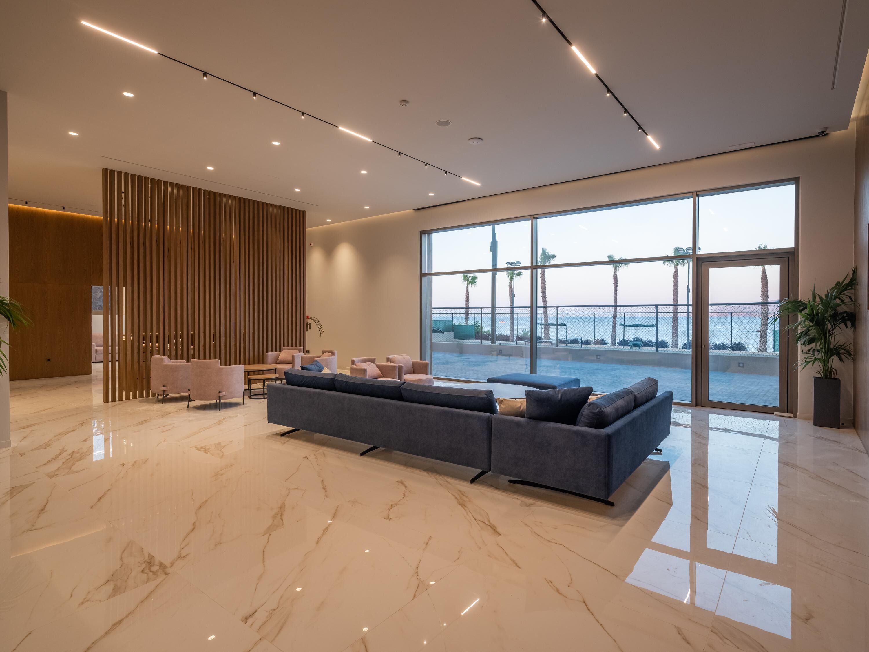 A modern lobby with a large seating area, a wooden partition, and a view of the sea through large windows.