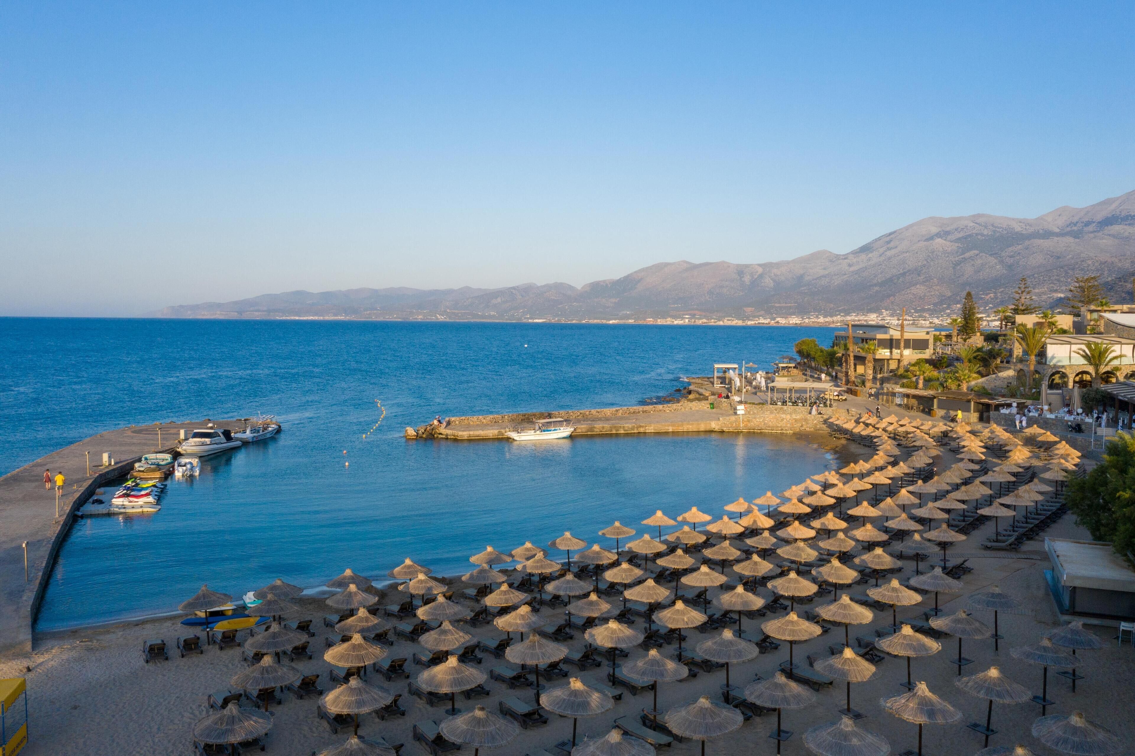 A coastal beach with umbrellas, a dock with boats, and mountains in the background.