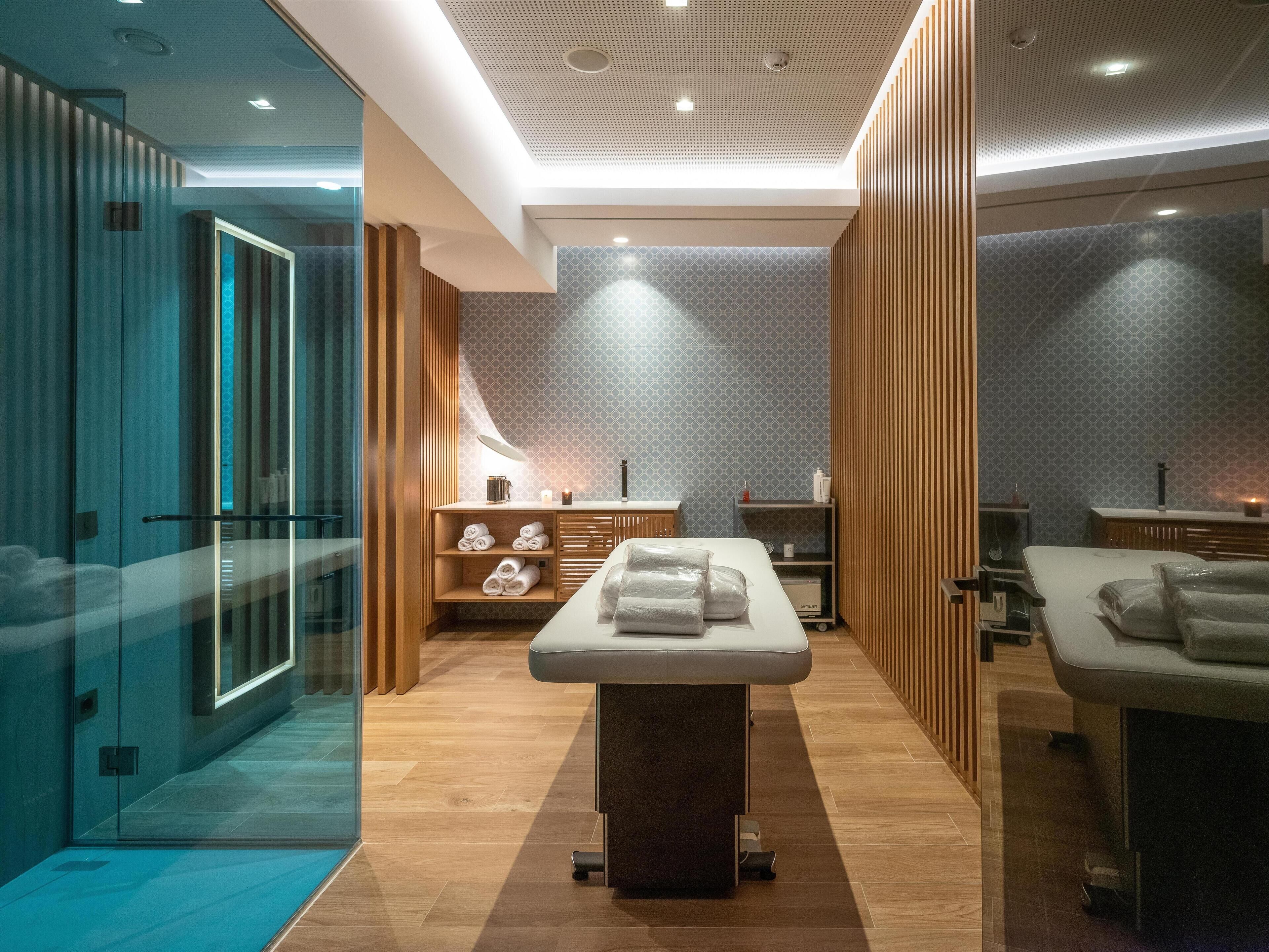A modern spa room with a massage table, wooden shelving, and a glass shower area.