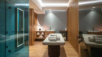 A modern spa room with a massage table, wooden shelving, and a glass shower area.