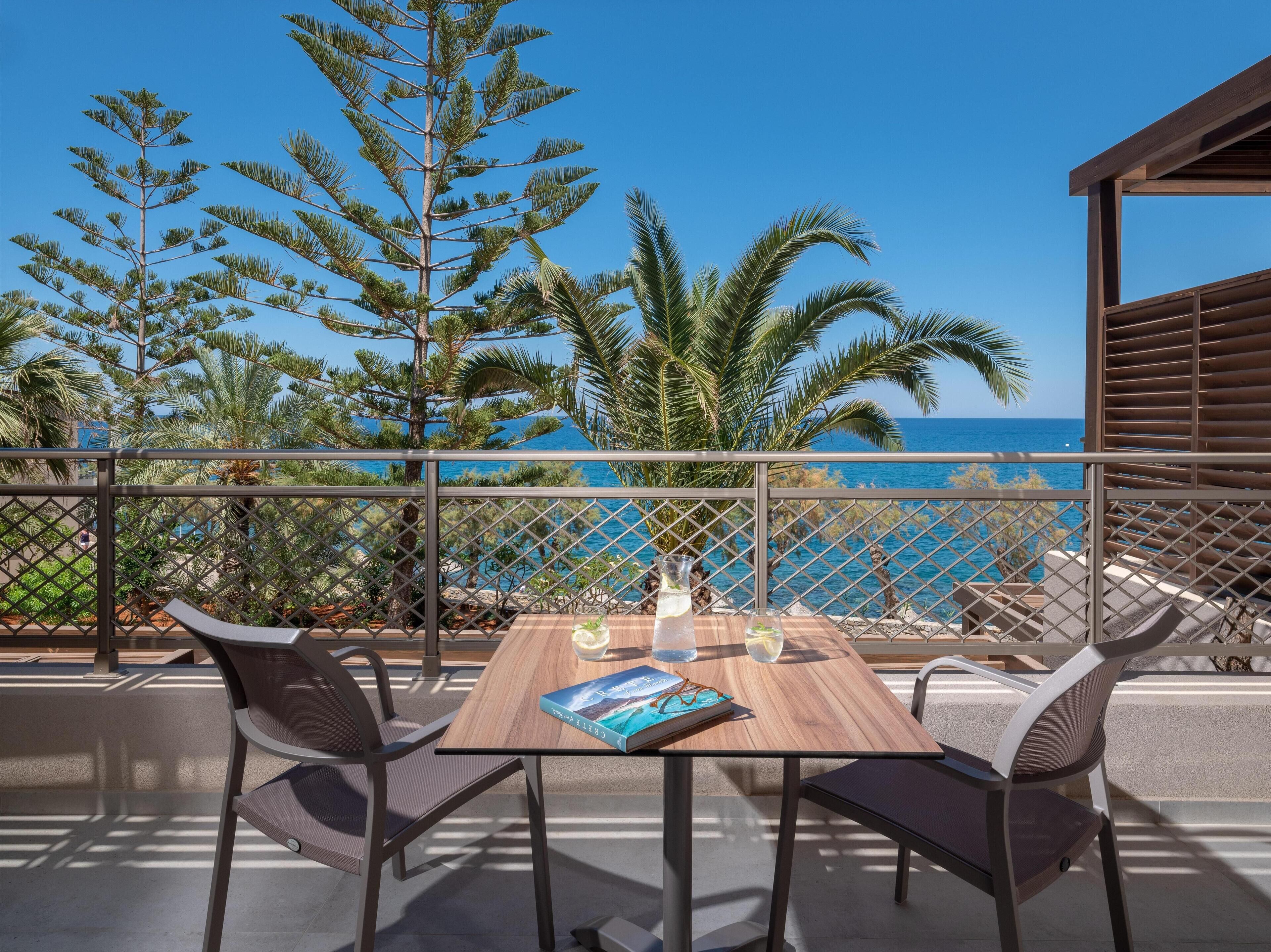 A wooden table with two chairs on a balcony overlooking the sea, with palm trees and a clear sky in the background.