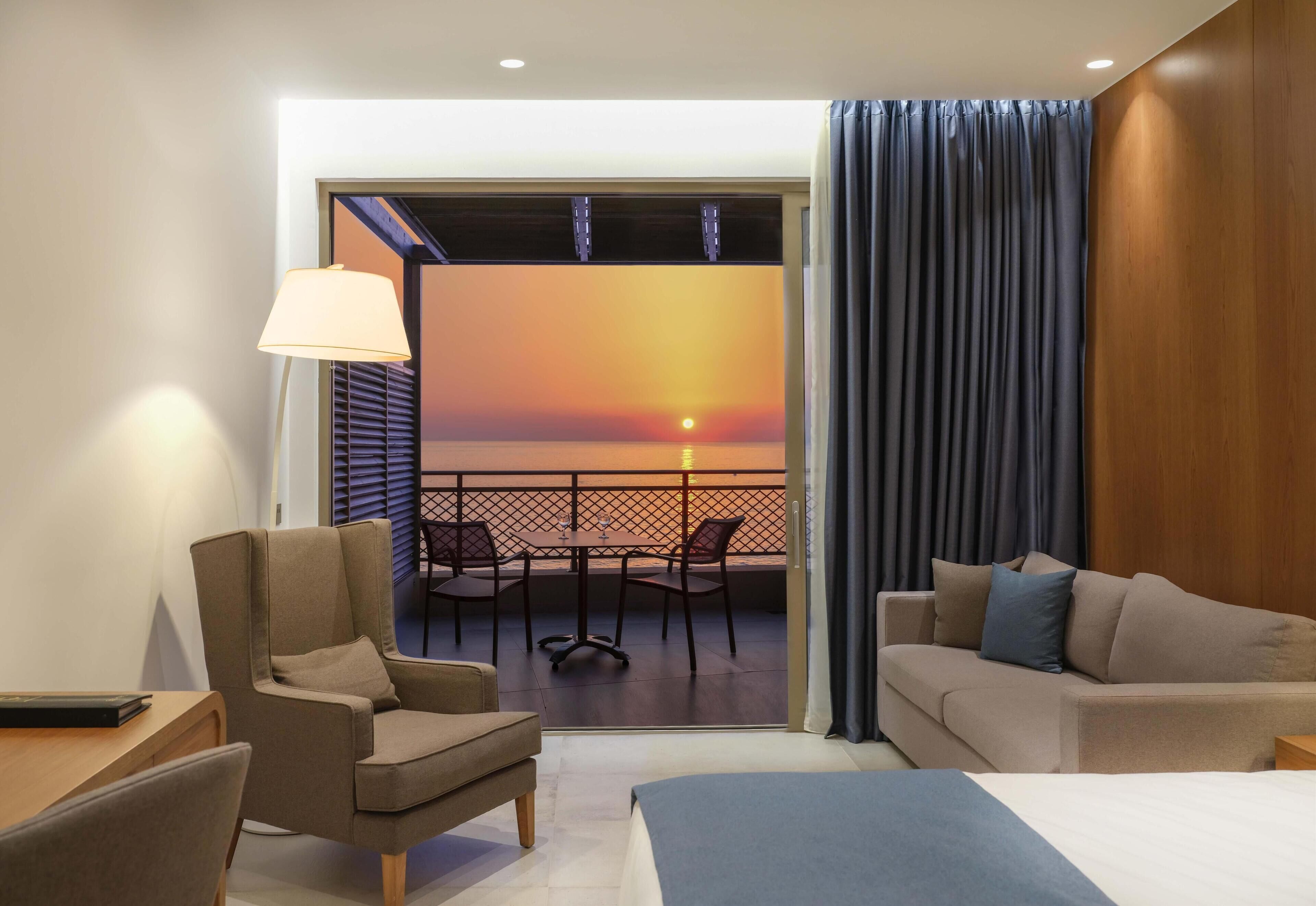 Habitación de hotel con cama, sofá, escritorio y silla. Tiene un balcón con vista al atardecer.
