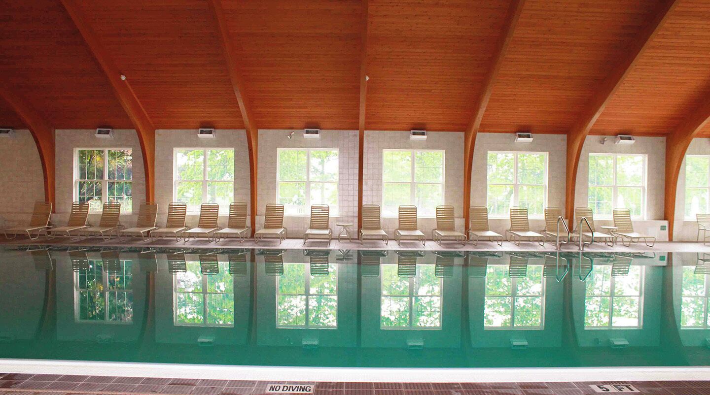 Una piscina interior con vigas de madera en el techo, varias ventanas y sillones de descanso a lo largo de la piscina.
