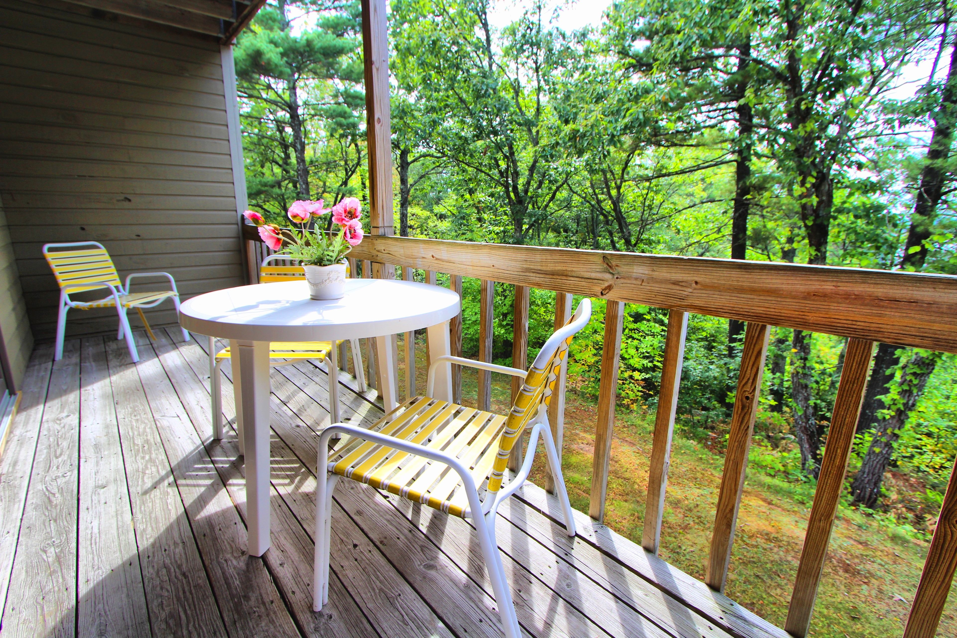 A wooden deck with a round table and two chairs, overlooking a forested area.