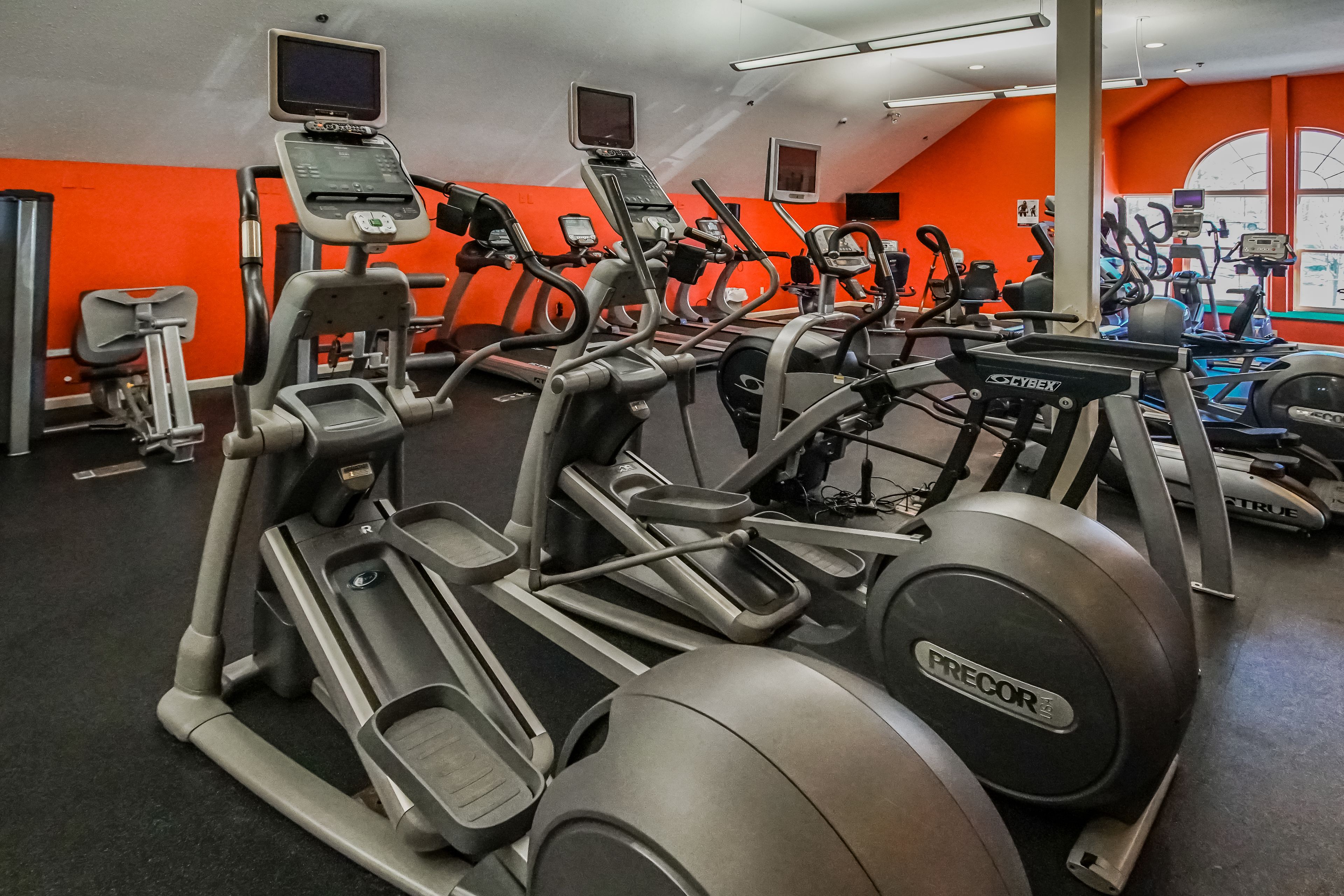 A gym with multiple treadmills and exercise bikes, bright orange walls, and a large window.
