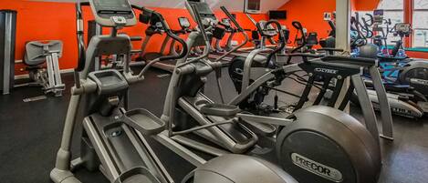 A gym with multiple treadmills and exercise bikes, bright orange walls, and a large window.