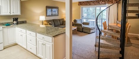 A kitchen with white cabinets, a granite countertop, and a coffee maker. There is a living area with a couch, a lamp, and a window with a view of trees. A staircase with wooden steps and a black railing leads to another level.