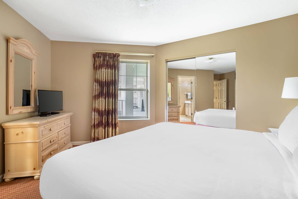 A hotel room with a large bed, a TV on a wooden dresser, a window with curtains, and a door leading to another room.