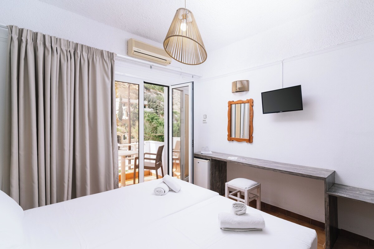 A hotel room with a bed, a desk, a chair, a television, and a sliding glass door to the outside.