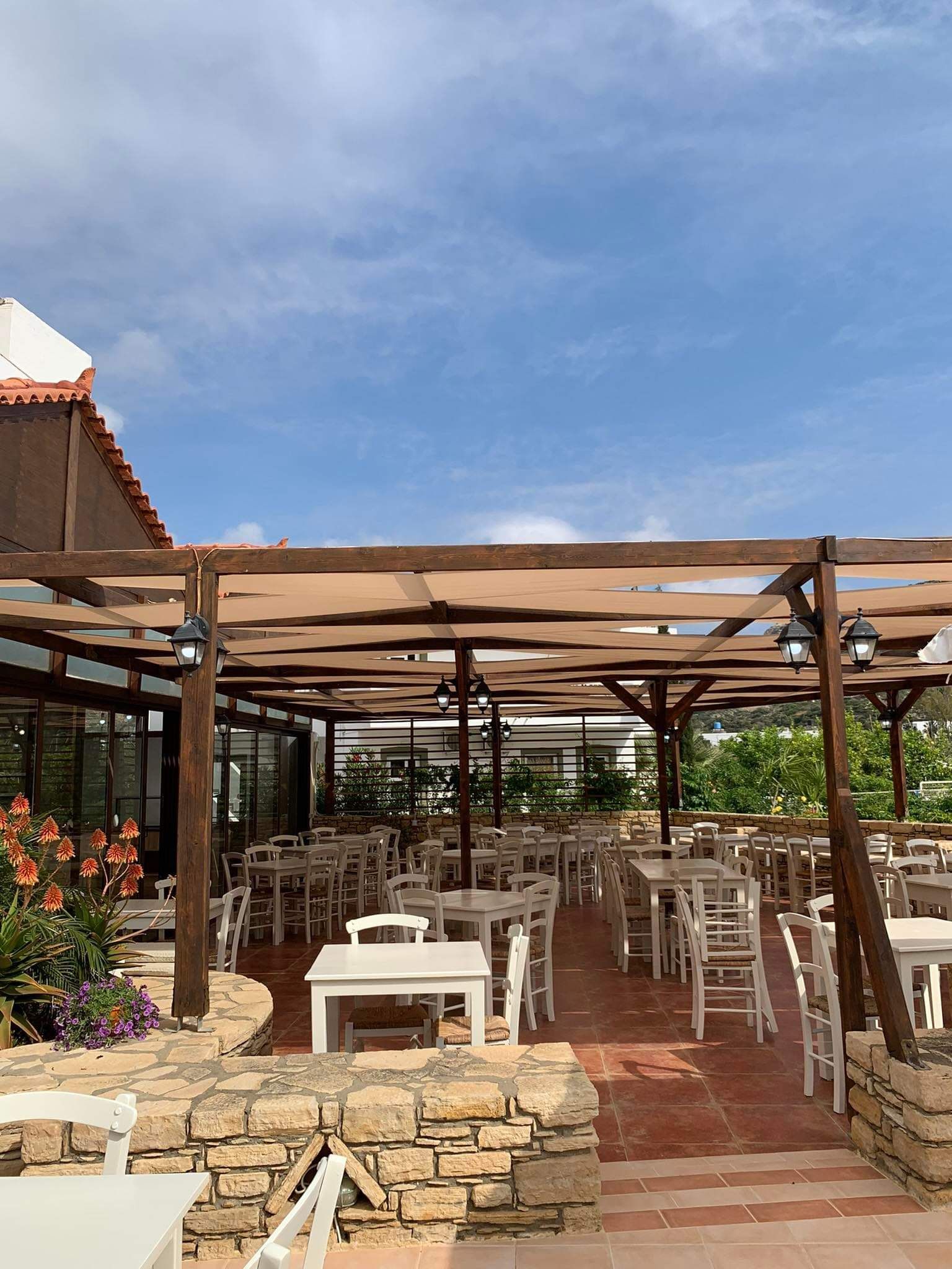 Un patio exterior con suelo de piedra, mesas y sillas blancas, y una estructura de pérgola de madera.