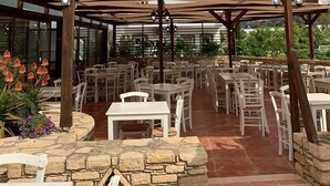 An outdoor patio with stone flooring, white tables and chairs, and a wooden pergola structure. - Elea Terra Hotel (Faistos)