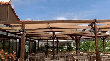 Un patio exterior con suelo de piedra, mesas y sillas blancas, y una estructura de pérgola de madera.