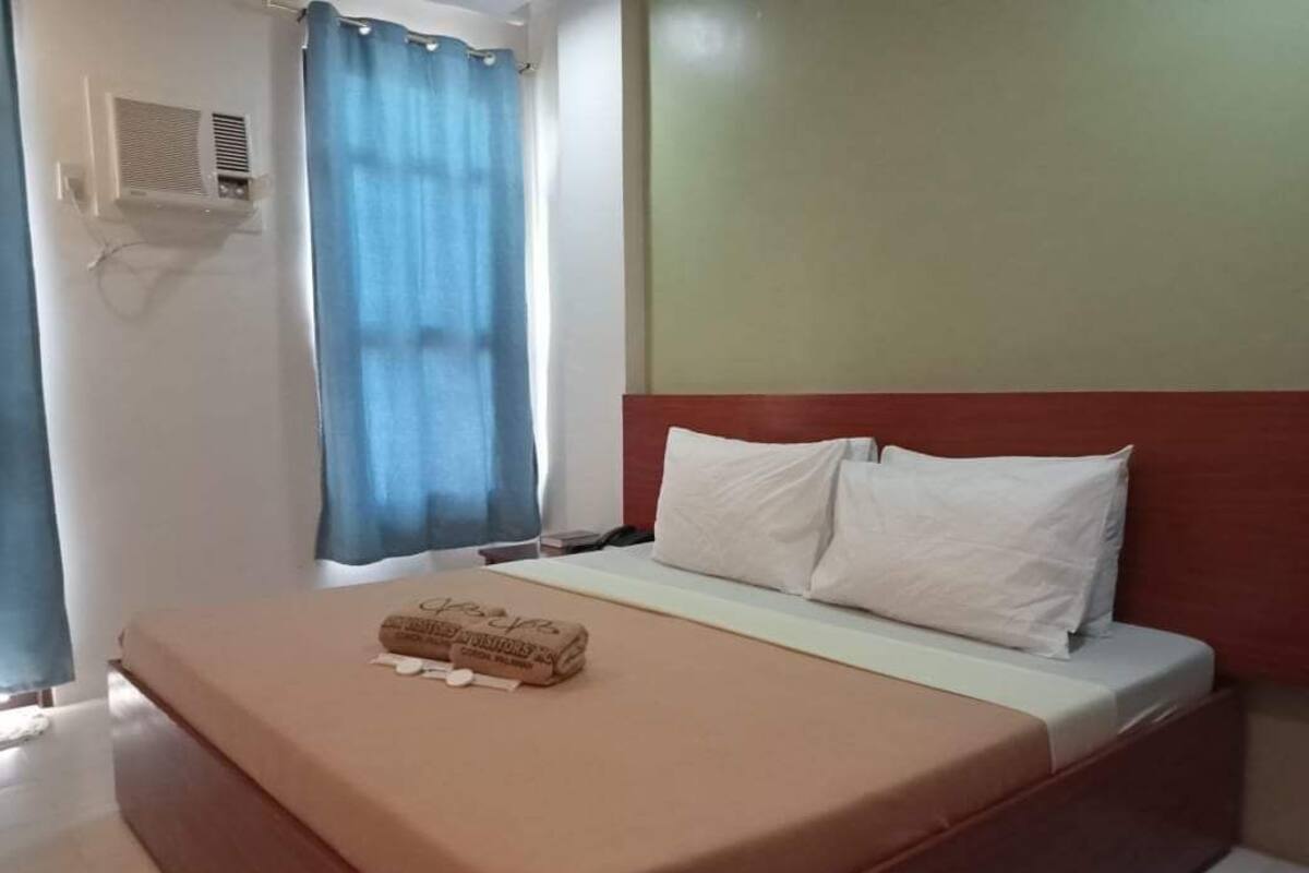 A neatly made bed with a brown bedspread and two white pillows, a blue curtain, and a wall-mounted air conditioning unit.