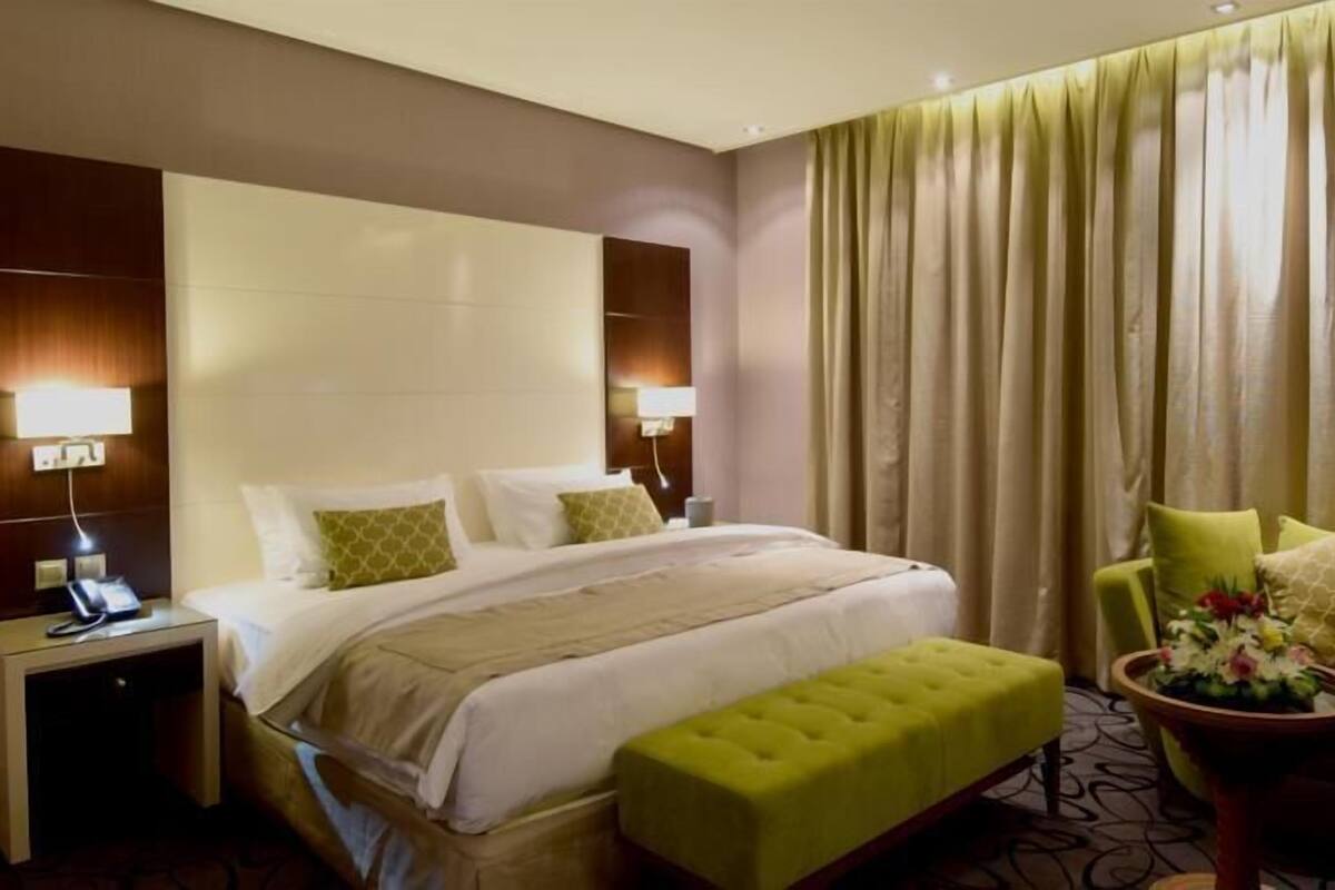 Une chambre d’hôtel avec un grand lit, un pouf vert, un fauteuil avec des coussins, une table de chevet avec un téléphone et une lampe fixée au mur.