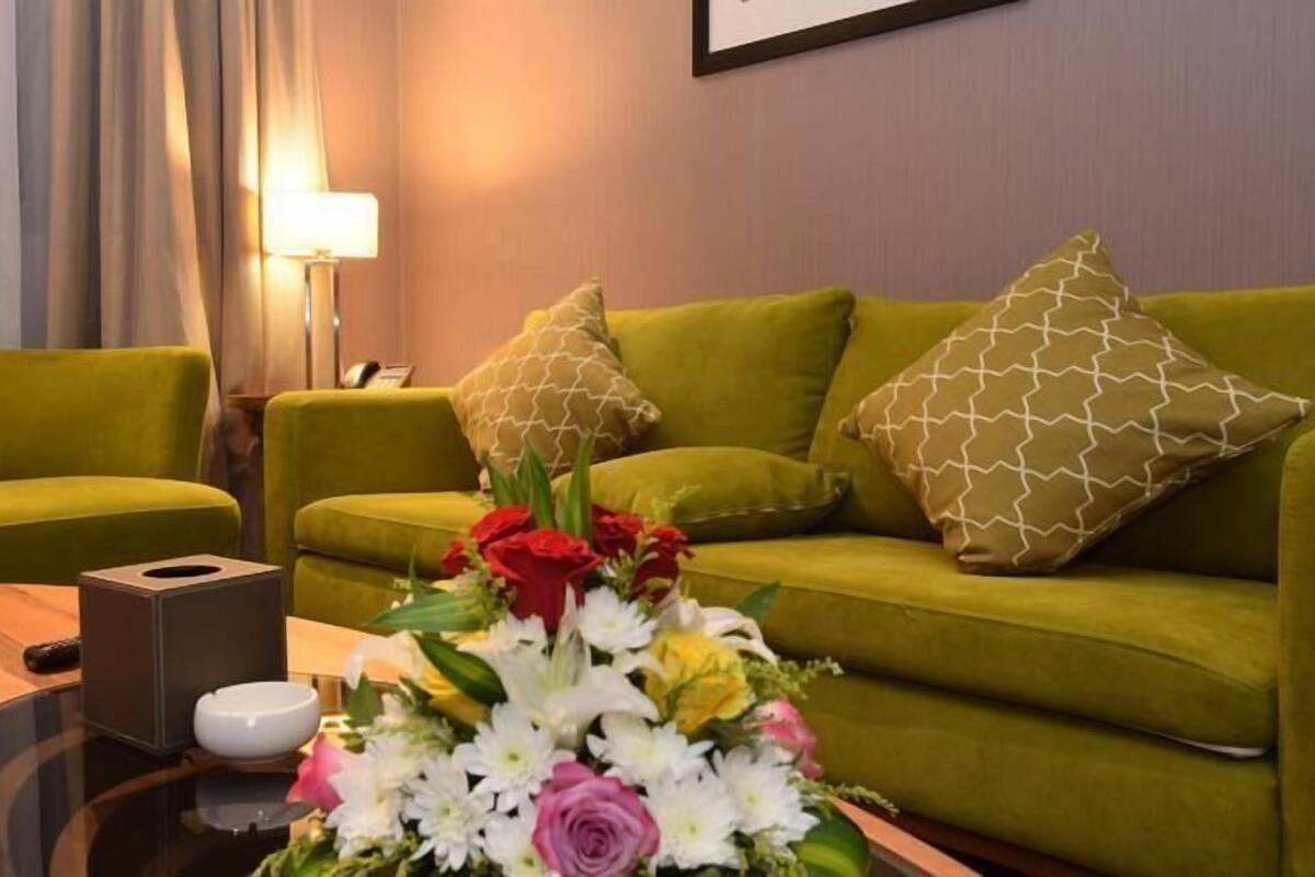 Une chambre d’hôtel avec un canapé vert, deux coussins assortis, une petite table sur laquelle se trouvent une boîte de mouchoirs et un bol, ainsi qu’un arrangement floral.
