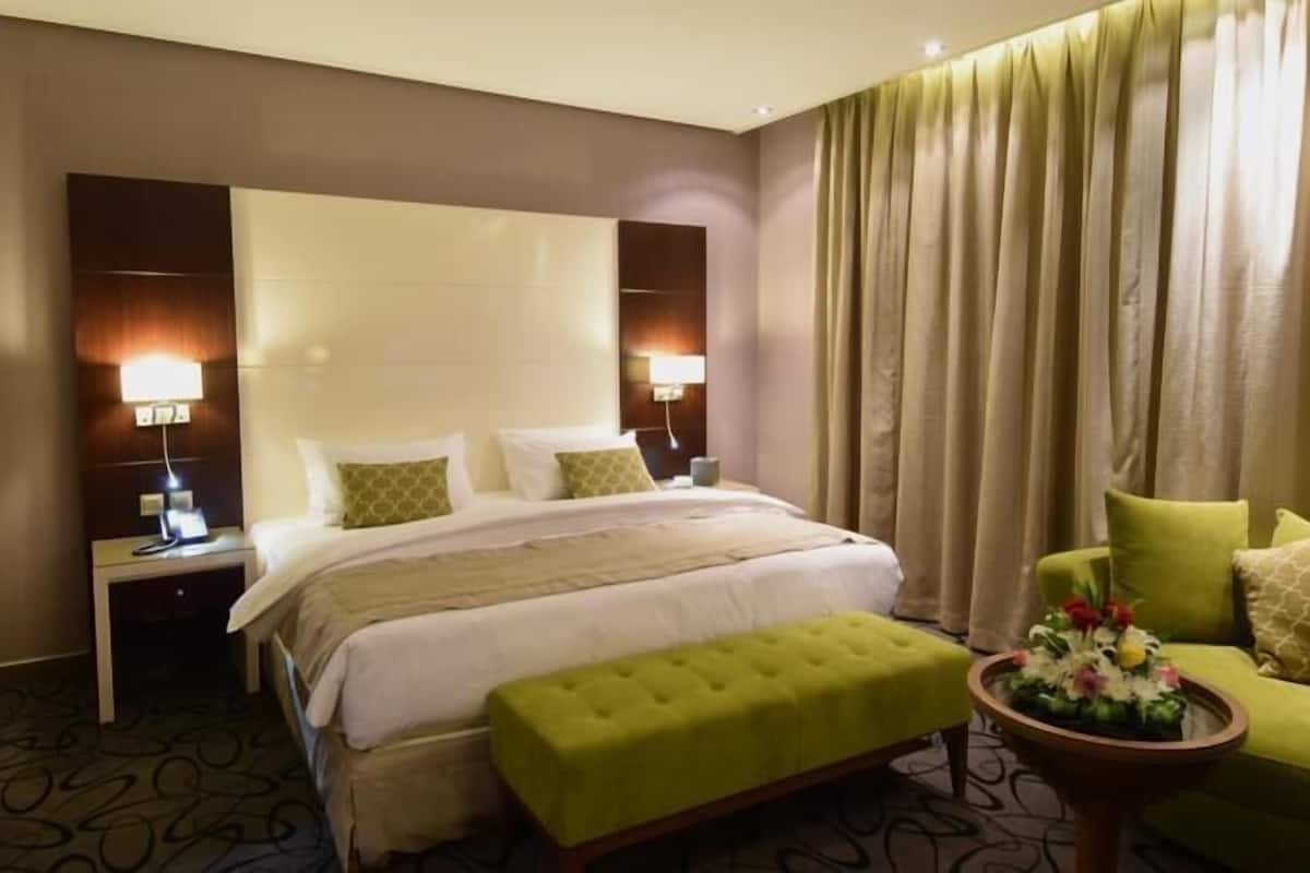 Une chambre d’hôtel avec un grand lit, deux lampes de chevet, un pouf vert, une petite table avec des fleurs et un fauteuil vert.