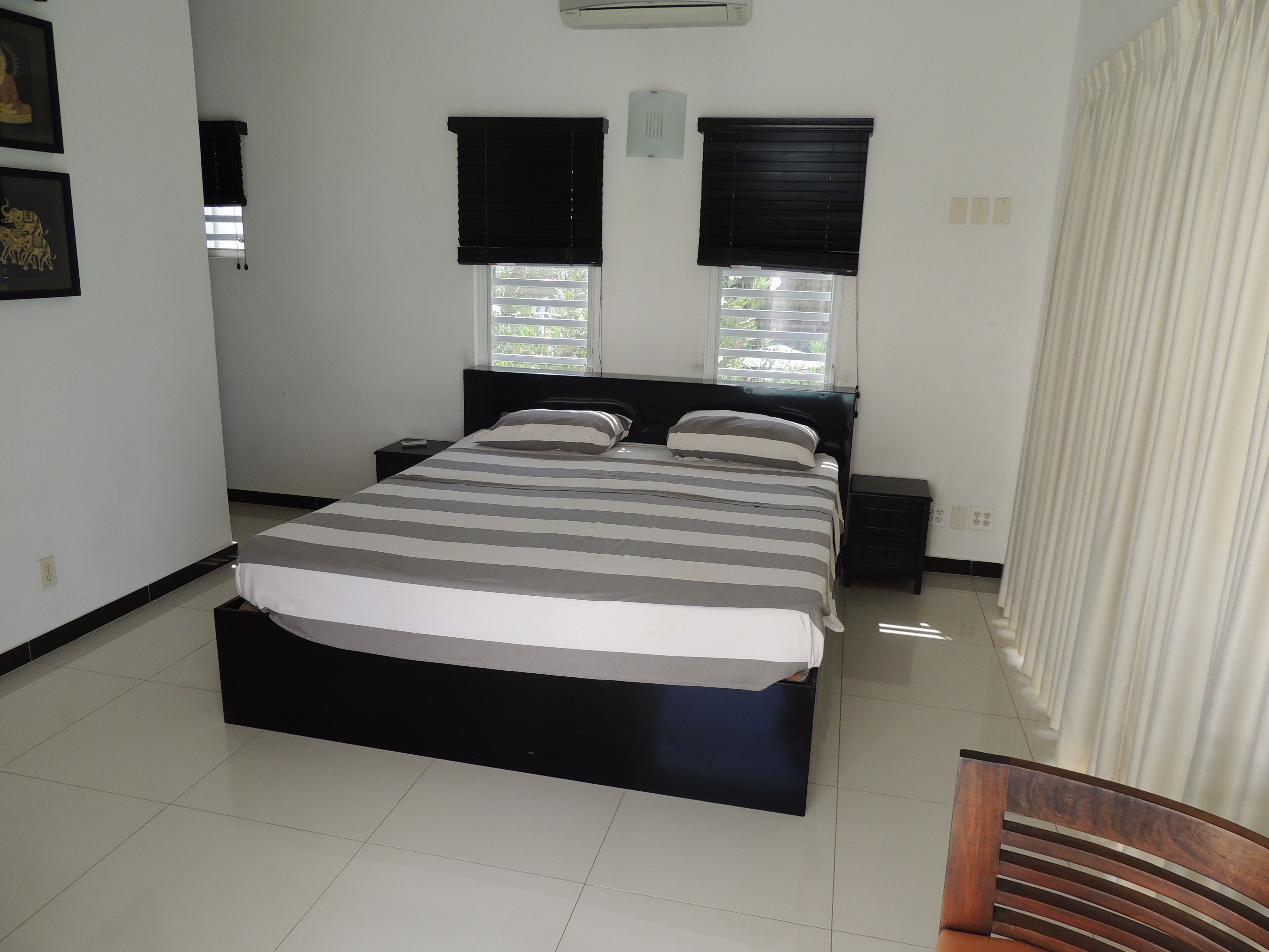 A bedroom with a large bed, two bedside tables, a window with blinds, and a curtain.