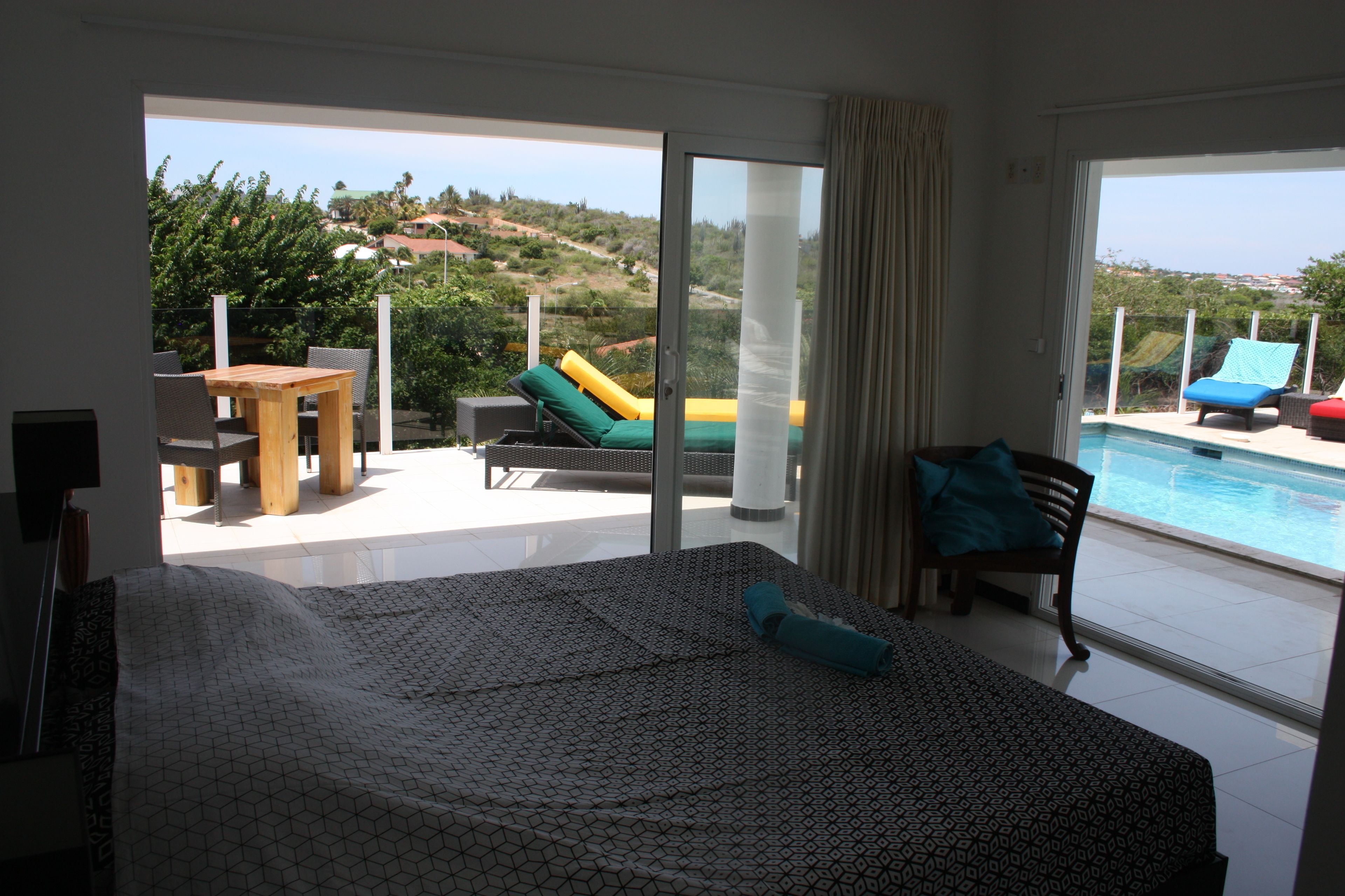 A hotel room with a bed, a table, and chairs, a balcony with a pool, and a view of the landscape.