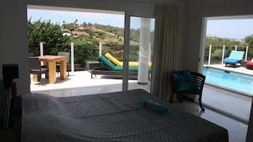 A hotel room with a bed, a table, and chairs, a balcony with a pool, and a view of the landscape.