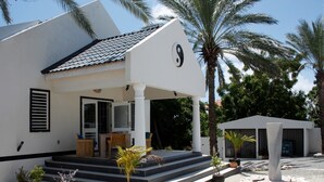 A modern building with a white exterior, a tiled roof, and a covered entrance area with steps leading up to it. There are potted plants and a large stone sculpture in the foreground. - Sun Sea Sleep Apartments (Willemstad)