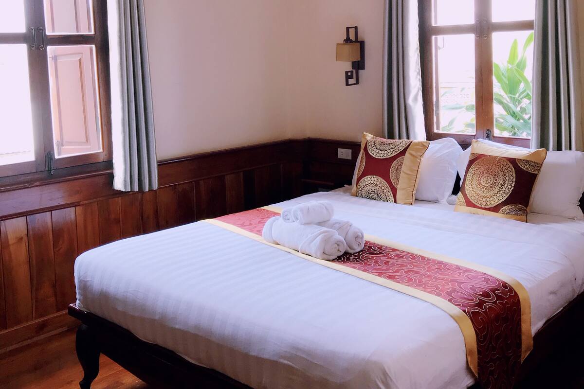 A hotel room with a large bed, wooden headboard, and two windows with curtains.